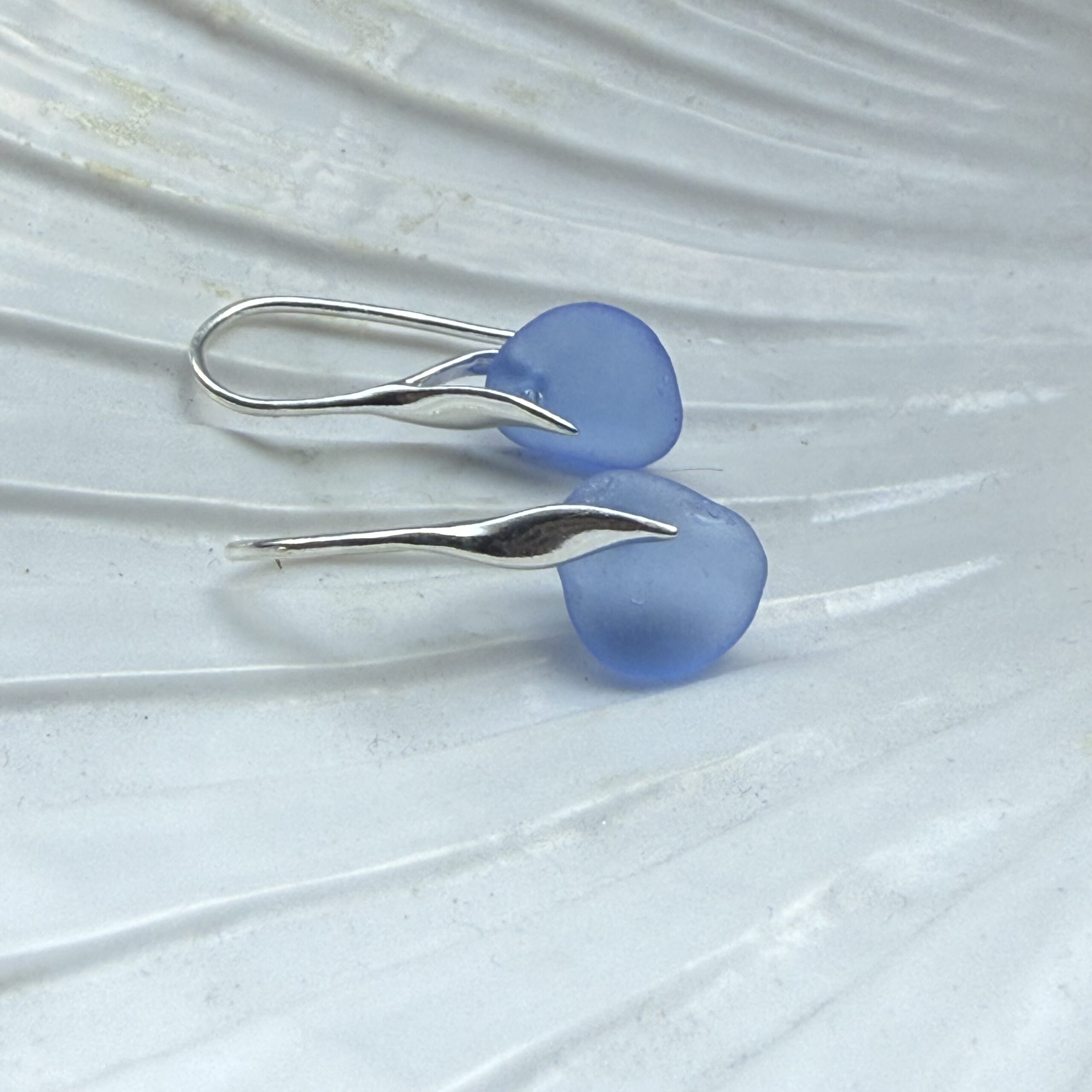 blauwe zeeglas oorbellen met zilveren haakjes liggend op een schelp