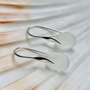 Witte zeeglas oorbellen met een zilveren oorhaakjes liggend op een schelp