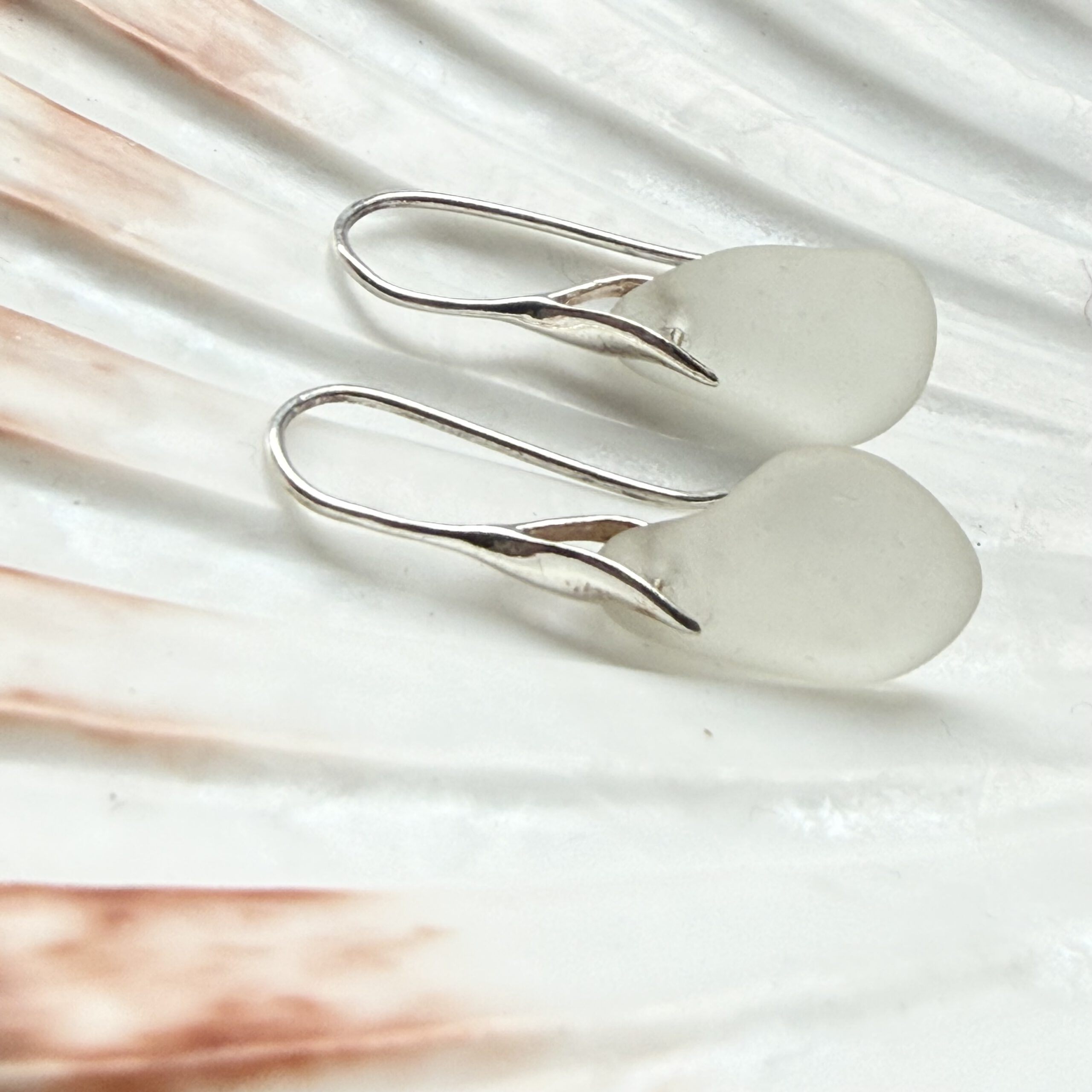 witte zeeglas oorbellen met zilveren oorhaakjes liggend op een schelp
